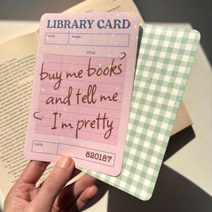 May include: A pink library card with the text "buy me books and tell me I'm pretty" and a green gingham bookmark. The card has a glittery finish and the number 820187.