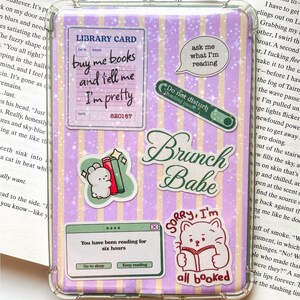 May include: A clear protective case for an e-reader or tablet featuring a purple and yellow vertical striped background with a sparkly star pattern. The case is decorated with various stickers including a library card, a cat reading a book, and text.