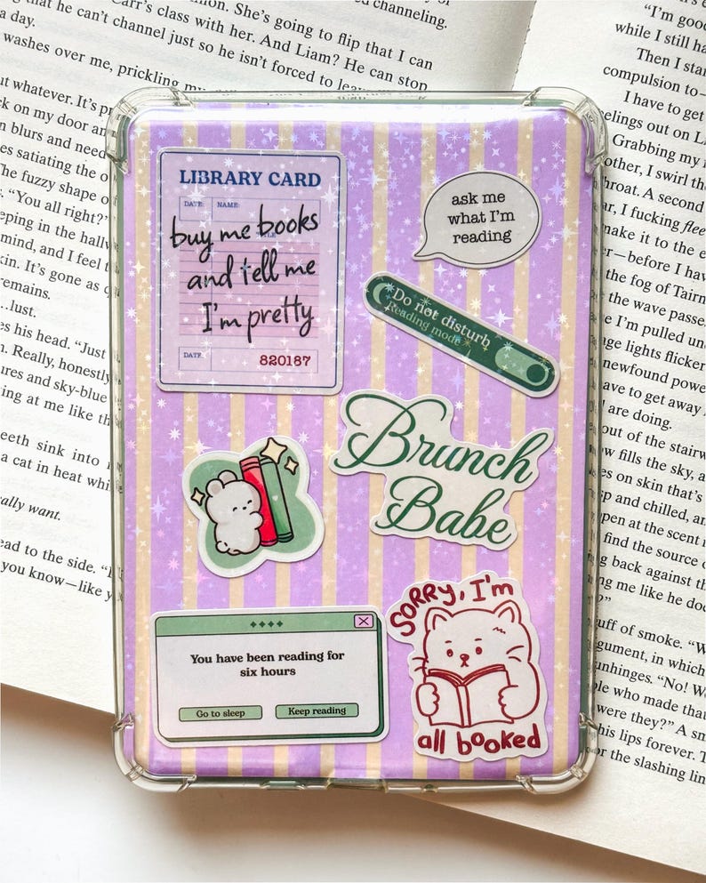 May include: A clear protective case for an e-reader or tablet featuring a purple and yellow vertical striped background with a sparkly star pattern. The case is decorated with various stickers including a library card, a cat reading a book, and text.