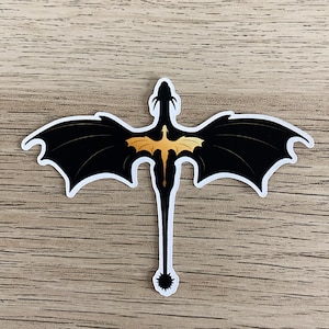 Peut inclure: Un autocollant d&eacute;coup&eacute; repr&eacute;sentant la silhouette noire d'un dragon aux grandes ailes d&eacute;ploy&eacute;es et &agrave; la longue queue termin&eacute;e par une nageoire &eacute;pineuse. Au centre du dos du dragon se trouve un petit motif de dragon dor&eacute; aux ailes d&eacute;ploy&eacute;es.