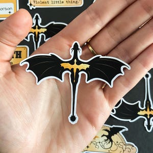 Peut inclure: Un autocollant d&eacute;coup&eacute; repr&eacute;sentant un dragon noir stylis&eacute; avec de grandes ailes pointues et une longue queue fine se terminant par une boule &agrave; pointes. Le dragon porte une silhouette de dragon jaune dor&eacute; plus petite sur le dos.
