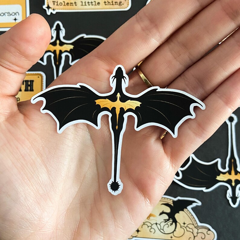 Peut inclure: Un autocollant d&eacute;coup&eacute; repr&eacute;sentant un dragon noir stylis&eacute; avec de grandes ailes pointues et une longue queue fine se terminant par une boule &agrave; pointes. Le dragon porte une silhouette de dragon jaune dor&eacute; plus petite sur le dos.