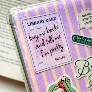 May include: A clear protective case featuring a light purple and yellow striped background with star patterns. It displays a library card sticker with the text: buy me books and tell me I'm pretty, alongside other stickers including a small bear.