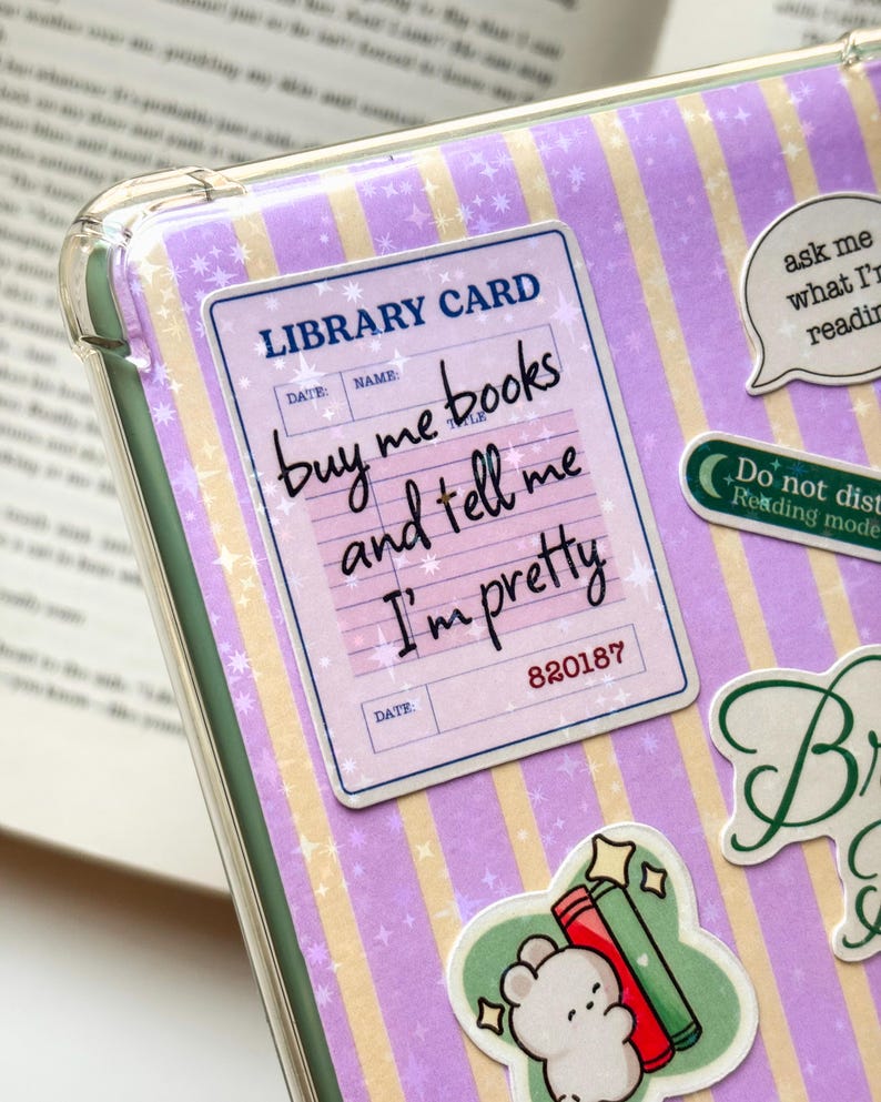 May include: A clear protective case featuring a light purple and yellow striped background with star patterns. It displays a library card sticker with the text: buy me books and tell me I'm pretty, alongside other stickers including a small bear.