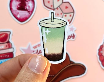 May include: A die-cut sticker featuring a beverage in a clear plastic cup with a lid and straw. The drink has a gradient color scheme transitioning from a dark brown base to a light green top, accented with white highlights to suggest a glossy finish.