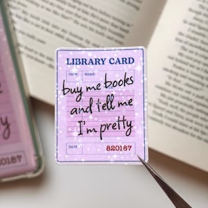 May include: A rectangular sticker designed as a vintage library card with a light purple background and white star accents. The text reads LIBRARY CARD at the top and buy me books and tell me I'm pretty in black cursive script, with 820187 at the bottom.