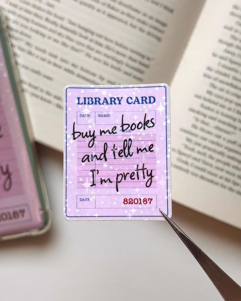 May include: A rectangular sticker designed as a vintage library card with a light purple background and white star accents. The text reads LIBRARY CARD at the top and buy me books and tell me I'm pretty in black cursive script, with 820187 at the bottom.