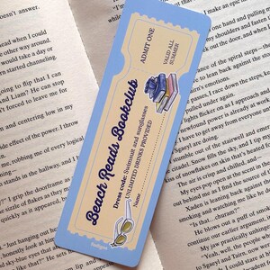 Pode incluir: Um marcador de página retangular desenhado como um bilhete vintage com uma borda azul clara e um centro creme. Apresenta o texto Beach Reads Bookclub, ADMIT ONE, VALID ALL SUMMER e UNLIMITED DRINKS PROVIDED, com ilustrações de livros empilhados e óculos de sol.