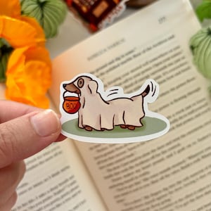 May include: A Halloween sticker showing a cartoon dog dressed as a ghost, carrying an orange bucket with "BOO" on it. The sticker has a white background and a green border, perfect for spooky season.