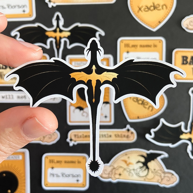Peut inclure: Un autocollant d&eacute;coup&eacute; repr&eacute;sentant la silhouette d'un dragon noir avec un embl&egrave;me de dragon dor&eacute; centr&eacute; sur son dos. Le dragon poss&egrave;de de grandes ailes pointues et une longue queue fine se terminant par une pointe circulaire dentel&eacute;e.