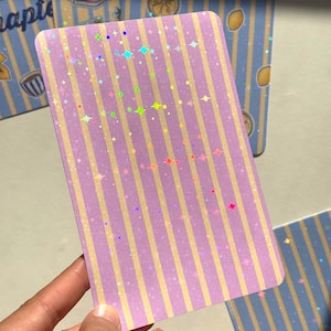 May include: A rectangular card featuring a vertical striped pattern in shades of light purple and pale yellow. The surface is covered in a holographic finish that displays small, scattered star and sparkle shapes in various rainbow colors.