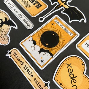Peut inclure: Une collection d'autocollants comprenant un panneau rectangulaire jaune Basgiath War College, un dragon noir, une carte jaune marqu&eacute;e Fantasy Reader avec un soleil noir, une banni&egrave;re jaune avec le texte Violent little thing et un c&oelig;ur jaune marqu&eacute; Xaden.