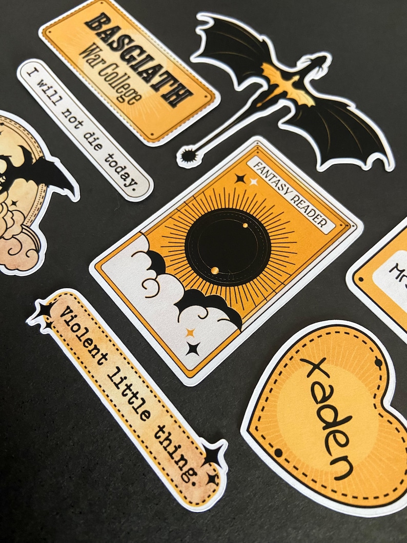 Peut inclure: Une collection d'autocollants comprenant un panneau rectangulaire jaune Basgiath War College, un dragon noir, une carte jaune marqu&eacute;e Fantasy Reader avec un soleil noir, une banni&egrave;re jaune avec le texte Violent little thing et un c&oelig;ur jaune marqu&eacute; Xaden.