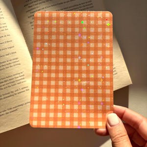 Op de afbeelding: Een rechthoekige kaart met een oranje en wit gingham ruitpatroon. Het oppervlak van de kaart is bedekt met een holografische afwerking die kleine, veelkleurige stervormige glitters over het gehele ruitpatroon weergeeft.