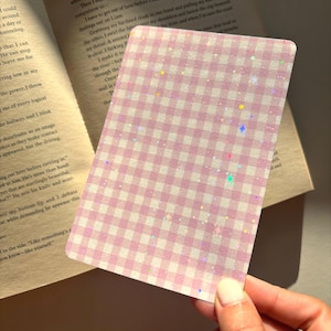 Könnte beinhalten: Eine rechteckige Karte mit einem hellrosa-weißen Vichy-Karo-Muster. Die Oberfläche der Karte ist mit einem holografischen Finish versehen, das kleine, mehrfarbige sternförmige Glitzerpunkte über das gesamte Karomuster hinweg zeigt.