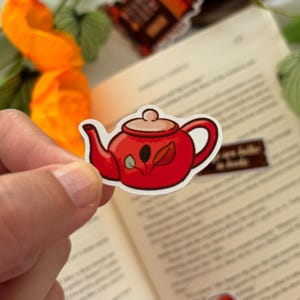 Peut inclure: Autocollant de théière rouge avec bordure blanche et motif de feuilles. L'autocollant est tenu devant un livre ouvert et d'autres autocollants. La théière a un bec et une anse incurvés.