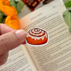 以下が含まれることがあります： 白いアイシングの渦巻きがのった茶色のシナモンロールのダイカットステッカー。イラストは、渦巻き状の中心部と、側面を伝って垂れる厚い白いグレーズがかかった、黄金色のペストリーを描いています。縁は細い白枠で囲まれています。
