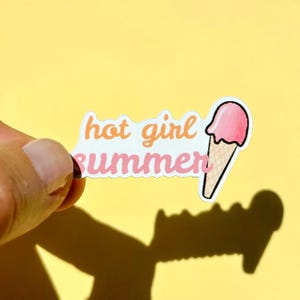 May include: A die-cut sticker featuring the text "hot girl summer" in orange and pink cursive lettering, accompanied by a graphic of a pink ice cream scoop in a tan waffle cone. The sticker has a white border and a matte finish.