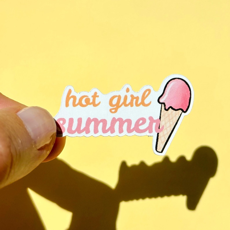 May include: A die-cut sticker featuring the text "hot girl summer" in orange and pink cursive lettering, accompanied by a graphic of a pink ice cream scoop in a tan waffle cone. The sticker has a white border and a matte finish.