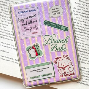 May include: A clear protective case for an electronic device featuring a purple and yellow striped background with a star pattern. The case is decorated with various stickers including a library card, a cat reading a book, and text reading Brunch Babe.