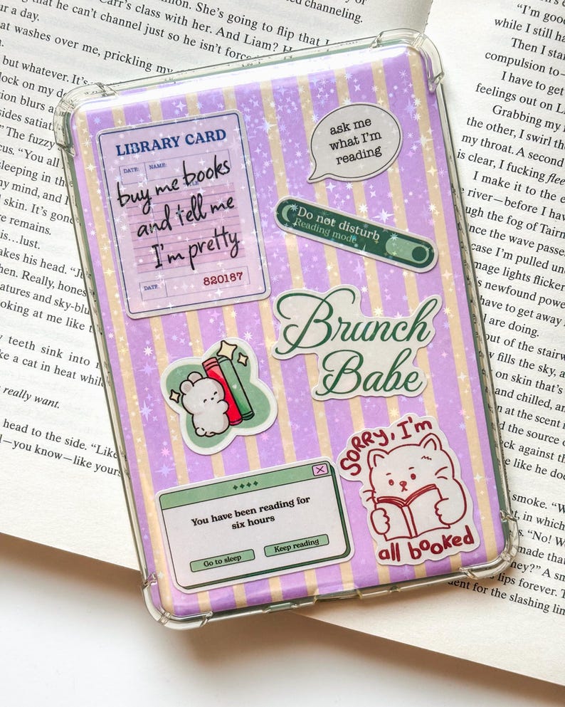 May include: A clear protective case for an electronic device featuring a purple and yellow striped background with a star pattern. The case is decorated with various stickers including a library card, a cat reading a book, and text reading Brunch Babe.