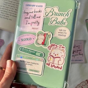 May include: A light green e-reader case decorated with various stickers including a library card reading buy me books and tell me I'm pretty, text saying Brunch Babe, a cat reading a book with text sorry I'm all booked, and a reading mode toggle.