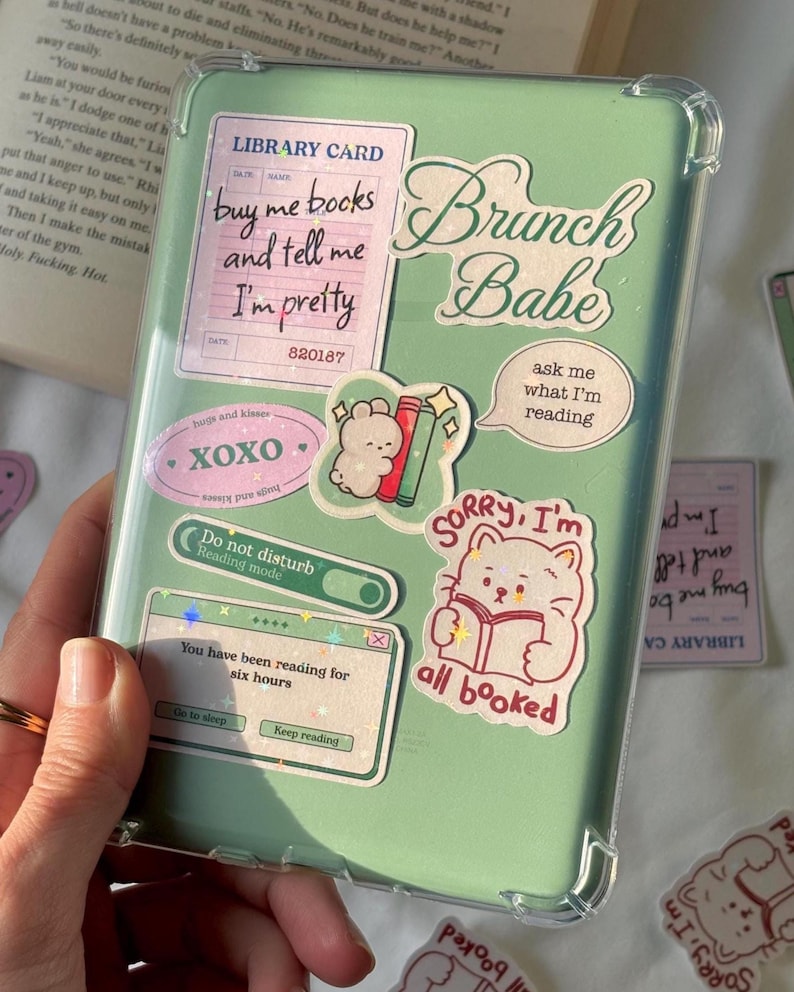 May include: A light green e-reader case decorated with various stickers including a library card reading buy me books and tell me I'm pretty, text saying Brunch Babe, a cat reading a book with text sorry I'm all booked, and a reading mode toggle.