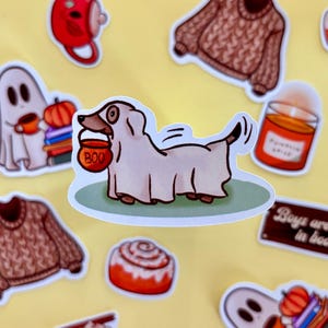 May include: A selection of autumn-themed stickers on a yellow backdrop. The stickers feature a dog in a ghost costume with a "BOO" candy bucket, a brown jumper, a cinnamon swirl, a candle, and a ghost.