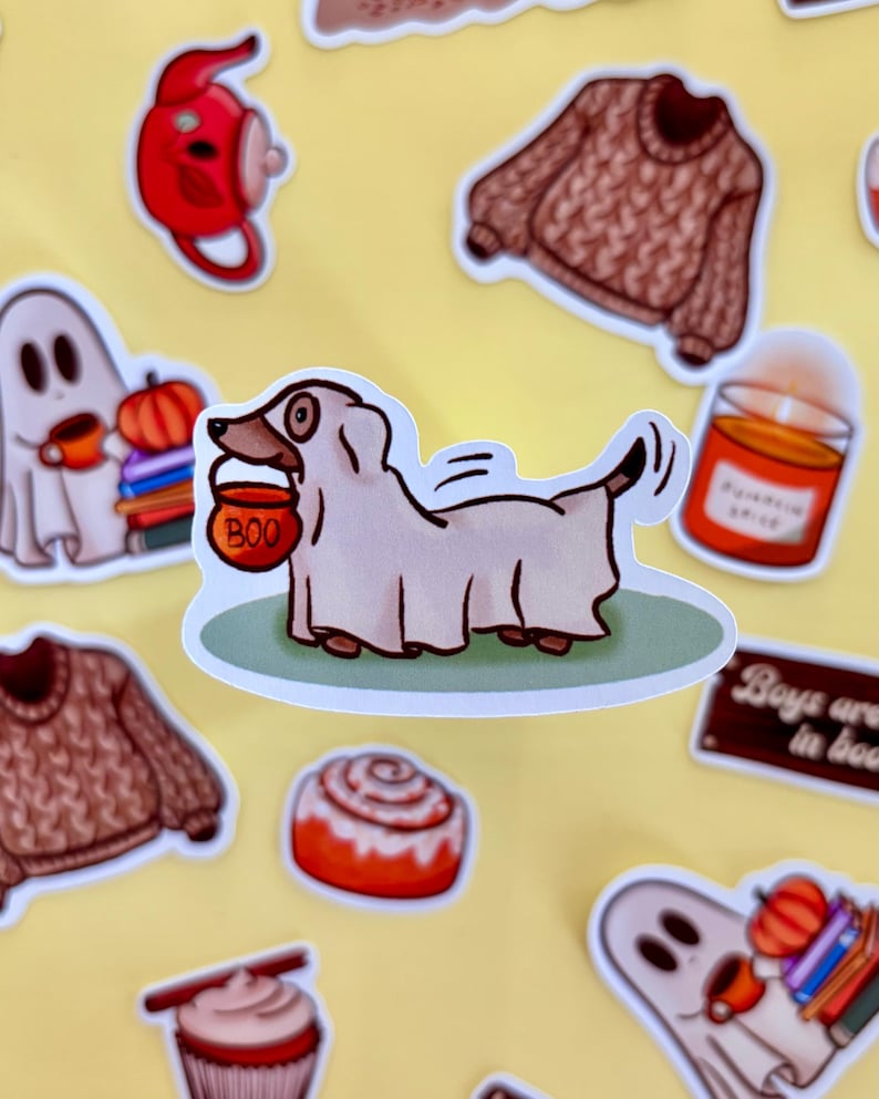 May include: A selection of autumn-themed stickers on a yellow backdrop. The stickers feature a dog in a ghost costume with a "BOO" candy bucket, a brown jumper, a cinnamon swirl, a candle, and a ghost.