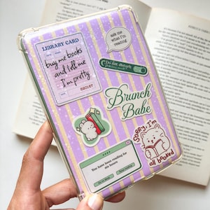 May include: A clear e-reader case featuring a purple and yellow striped background with a star pattern. The case is decorated with various stickers, including a library card design, a speech bubble, a cat reading a book, and text boxes.