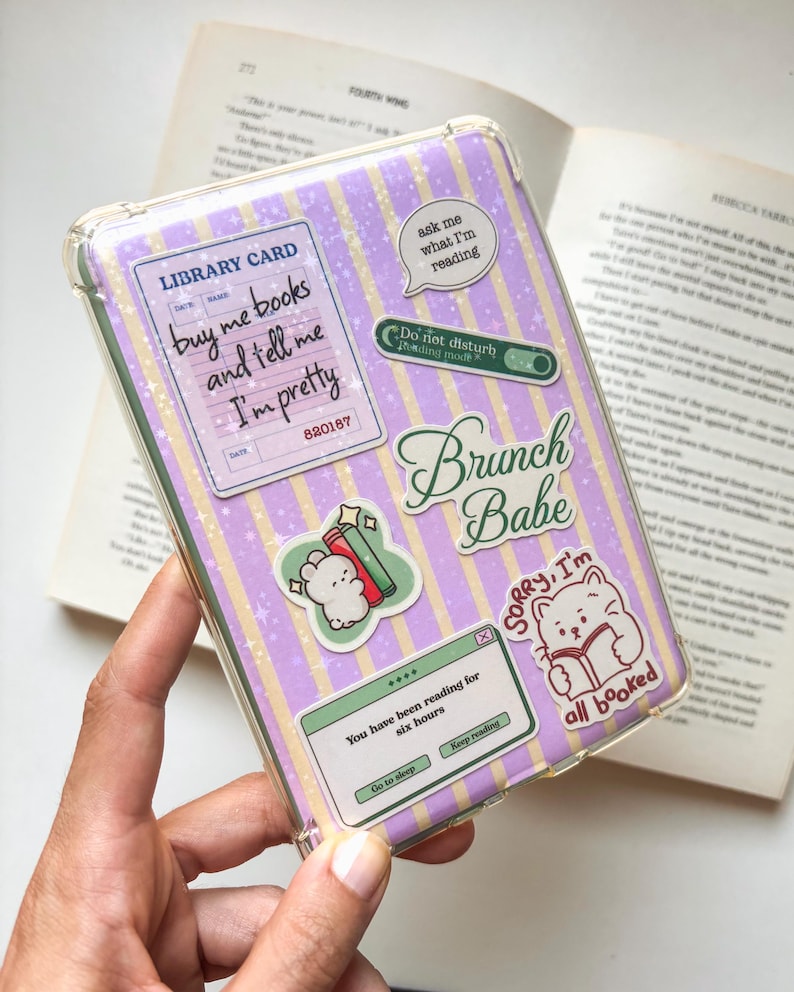May include: A clear e-reader case featuring a purple and yellow striped background with a star pattern. The case is decorated with various stickers, including a library card design, a speech bubble, a cat reading a book, and text boxes.