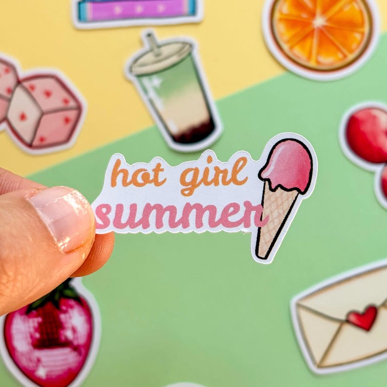 May include: A die-cut vinyl sticker featuring the text hot girl summer in orange and pink cursive lettering, accompanied by a graphic of a pink ice cream cone. The sticker is held against a background of various other colorful food-themed stickers.