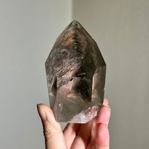May include: A clear quartz crystal point with a smoky grey and pink color variation. The crystal is faceted and has a pointed tip.