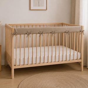 May include: A light wood crib featuring a rectangular beige fabric rail cover secured with six matching ribbon ties. The crib has vertical slats, a white mattress, and a clean frame design, providing a neutral and functional sleeping space.