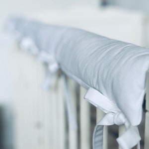 Puede incluir: Cojín de protección de barandilla de cuna blanco con lazos blancos. El cojín está hecho de tela suave y está diseñado para proteger a los bebés de golpes y moretones.