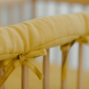 Puede incluir: Primer plano de un parachoques de cuna amarillo con un lazo atado en el extremo. El parachoques está hecho de una tela suave y texturizada y está diseñado para proteger a los bebés de golpes y moretones.