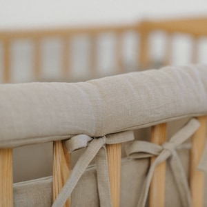 May include: Close-up of a beige linen crib bumper pad, tied to the wooden crib rails with matching fabric ties. The neutral color and natural texture create a soft, calming aesthetic for a baby's nursery.