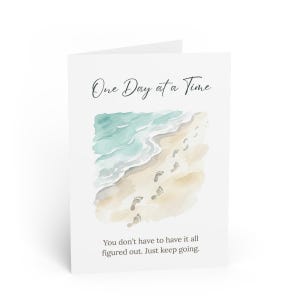 Peut inclure: Une carte de vœux blanche avec le texte "One Day at a Time" en écriture élégante. La carte présente une illustration à l'aquarelle d'une scène de plage avec des empreintes de pas dans le sable et l'océan. Le bas de la carte se lit : "You don't have to have it all figured out. Just keep going."
