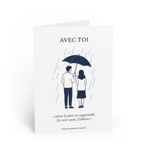 May include: White greeting card featuring the text "AVEC TOI" and a drawing of a couple sheltering under an umbrella in the rain. The card also includes the text "...même la pluie est supportable. (Le reste aussi, d'ailleurs.)" and "Joyeux anniversaire, mon abri."