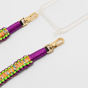 May include: A purple phone strap with a gold clasp and a colorful braided design. The strap is attached to a clear phone case.