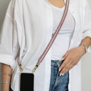 May include: A white button-down shirt with a pink and gold phone strap attached. The strap is worn diagonally across the chest and has a clear phone case attached to it.