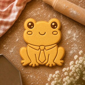 Könnte beinhalten: Ein froschförmiger Ausstecher mit einem lächelnden Gesicht und einer Krawatte. Der Ausstecher ist hellgelb und liegt auf einer mit Mehl bestäubten Holzoberfläche. Ein Nudelholz und ein rosa-weiß kariertes Tuch sind im Hintergrund.