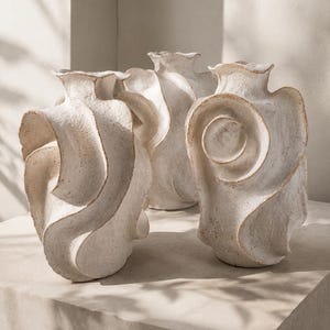 May include: Three off-white ceramic vases with a rough, textured surface and organic, swirling, wave-like forms. Each vase features a narrow neck and a wide, sculptural body with prominent, layered ridges that create a dynamic, fluid appearance.