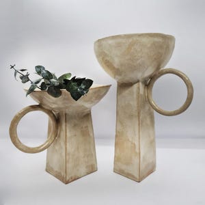 May include: Two beige ceramic vases with a unique design. Each vase has a wide, shallow bowl shape atop a rectangular base. One vase features a ring handle. The vases are displayed on a white surface, and one holds green leafy sprigs, showcasing a minimalist aesthetic.