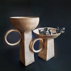 May include: Two beige ceramic pedestal bowls with circular handles. One bowl is empty, and the other contains dark gray stones and a sprig of greenery. The bowls have a textured surface and are set against a dark background.