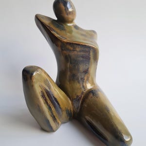 May include: A ceramic sculpture of a seated figure with an arm raised. The figure is glazed in shades of olive green, brown, and blue. The artwork has a smooth, glossy finish and is set against a plain white background.