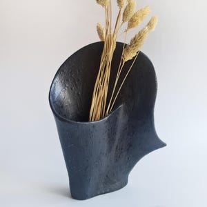 Puede incluir: Un jarrón de cerámica gris oscuro texturizado con una forma orgánica, que contiene un manojo de hierba ornamental beige seca. El jarrón tiene un diseño asimétrico único y se encuentra sobre un fondo blanco liso.