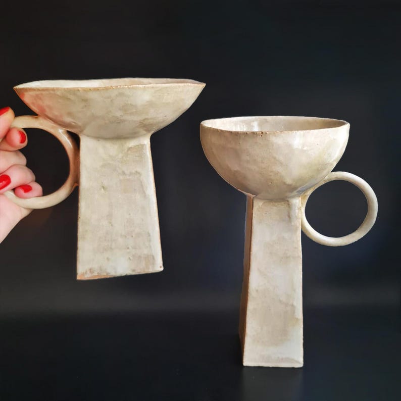 Puede incluir: Dos tazas de cer&aacute;mica blanquecina con un dise&ntilde;o &uacute;nico. Cada taza presenta una forma de cuenco ancho y poco profundo sobre una base rectangular. Un asa circular est&aacute; unida al lateral de cada taza. Las tazas tienen un acabado mate texturizado.