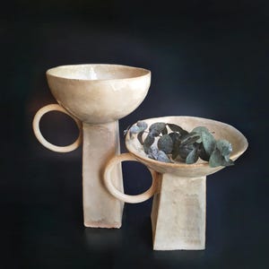 Puede incluir: Dos cuencos de pedestal de cer&aacute;mica blanquecina con asas circulares. Un cuenco contiene hojas verdes. Los cuencos tienen una superficie texturizada y est&aacute;n colocados sobre un fondo oscuro. Los cuencos son probablemente para fines decorativos.