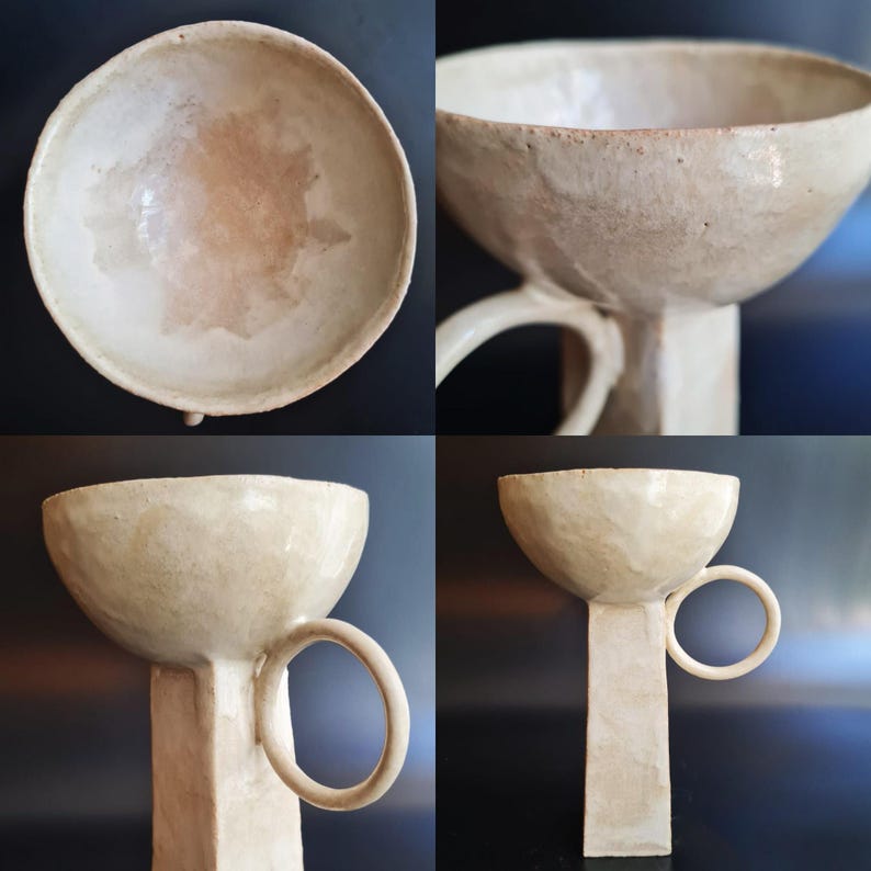 Puede incluir: Un cuenco de cer&aacute;mica blanco roto hecho a mano con un dise&ntilde;o &uacute;nico. El cuenco tiene una parte superior redondeada, una base cil&iacute;ndrica y un asa circular. La pieza de cer&aacute;mica tiene una superficie texturizada y una est&eacute;tica r&uacute;stica.